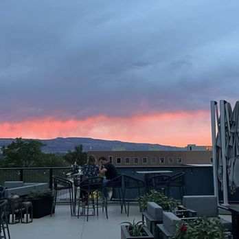 Devil's Kitchen - A rooftop dining space features tables, chairs, and umbrellas, with two people seated at a table engaged in conversation. The background shows a landscape with hills and a sky illuminated by a sunset, displaying shades of pink and orange amidst gray clouds. Potted plants are visible in the foreground, and the setting appears to be in an urban or suburban area. - New American Near Me - Grand Junction, Colorado