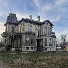 A large Victorian-style mansion built in the 19th century features ornate architectural details, tall arched windows, multiple chimneys, and a steeply pitched roof with decorative spires. The building is painted in light colors with darker trim, and a small staircase leads to the main entrance. Surrounding the mansion is a lawn with sparse patches of snow, and leafless trees are visible in the background, indicating a winter or early spring setting.
