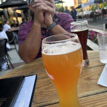 Martha's Exchange - A tall glass of beer with a foamy head is placed on a wooden table at an outdoor dining area, with a man in a striped shirt and cap in the background. - New American Near Me - Nashua, New Hampshire