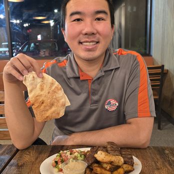 Armo Grill - A man with short black hair, wearing a gray polo shirt with orange accents and an 'I VOTED' sticker, is sitting at a restaurant table. He is smiling and holding a piece of flatbread in his right hand. In front of him is a white plate with grilled meats, potato wedges, a small salad, and a dipping sauce. The background shows a window with a nighttime scene outside, including parked cars and reflections of indoor lighting. - Armenian Near Me - Wheeling, Illinois