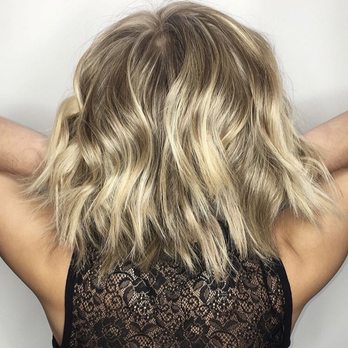 Pascal Pour Elle - A woman with shoulder-length, wavy blonde hair is shown from behind, wearing a black lace top, with arms raised and hands behind her head. - Hair Salons Near Me - Glencoe, Illinois