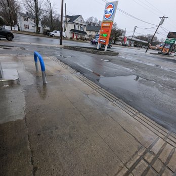 Dedham Gulf - Front display sign at the gas station - A wet parking lot outside a Gulf gas station on a rainy day, with puddles, a blue bike rack, and residential houses in the background. - Gas Stations Near Me - Dedham, Massachusetts
