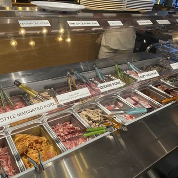 Boon Dee Moo Ka Ta - Meat - A buffet with metal trays containing different types of raw meats such as marinated pork, Thai style pork, and beef intestine, with labels and tongs for serving. - Thai Near Me - Flushing, New York