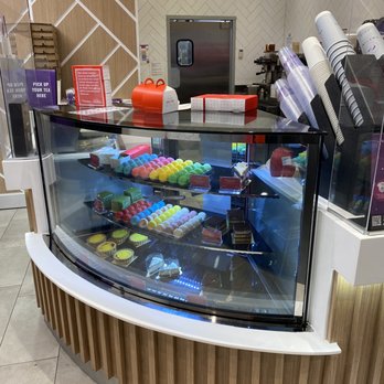 Chatime - Chatime - pastry display - The image features a curved glass display case filled with various desserts such as macarons in multiple colors and small tarts with yellow filling. The display is part of a cafe or bakery counter, with a wooden panel base and a tiled wall behind it. Items like cups and packaging materials are visible on the counter, along with signs and a red cash register. The background includes a door and some equipment, indicating an indoor food service environment. - Bubble Tea Near Me - Toronto, Ontario