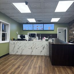 A casual restaurant interior features a marble counter with a white and gray veined pattern, digital menu screens mounted on a green wall behind the counter, and wood flooring. The ceiling has decorative panels with bright rectangular lights, and a window is visible on the left side. Equipment and supplies are placed on the counter, and a door is seen on the right side.