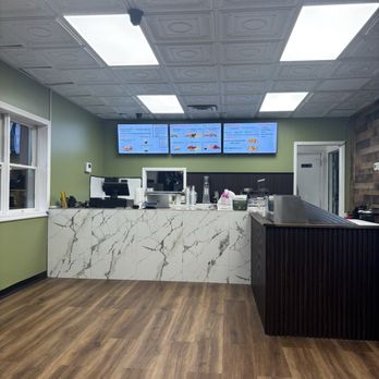 Sizzlings - A casual restaurant interior features a marble counter with a white and gray veined pattern, digital menu screens mounted on a green wall behind the counter, and wood flooring. The ceiling has decorative panels with bright rectangular lights, and a window is visible on the left side. Equipment and supplies are placed on the counter, and a door is seen on the right side. - Mediterranean Near Me - Oceanside, New York