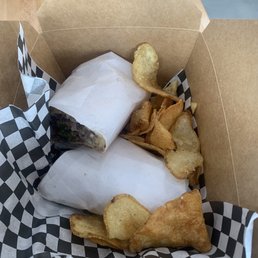 Steak & Cheese Deluxe - Two sandwiches wrapped in white paper are inside a checkered paper-lined takeout box, accompanied by potato chips.