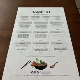 The menu - The menu from Bamboo by Kulantro is printed on a white sheet of paper placed on a wooden table. It lists various Vietnamese dishes including spring rolls, egg rolls, skewers, protein platter, bamboo bowl, bun bo hue, noodle bowl, simple pho, banh mi, rice super bowl, and vegan pho. Each item includes a description and price, with options for gluten-free and vegan modifications. The menu also features social media icons and a small illustration of a bowl of noodles with chopsticks and a lime.