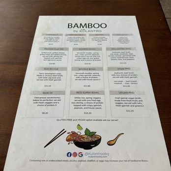 Bamboo by Kulantro - The menu - The menu from Bamboo by Kulantro is printed on a white sheet of paper placed on a wooden table. It lists various Vietnamese dishes including spring rolls, egg rolls, skewers, protein platter, bamboo bowl, bun bo hue, noodle bowl, simple pho, banh mi, rice super bowl, and vegan pho. Each item includes a description and price, with options for gluten-free and vegan modifications. The menu also features social media icons and a small illustration of a bowl of noodles with chopsticks and a lime. - Vietnamese Near Me - Albuquerque, New Mexico