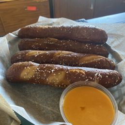 Pretzel sticks - Four pretzel sticks sprinkled with coarse salt are served with a cup of cheese sauce, on a paper-lined tray.