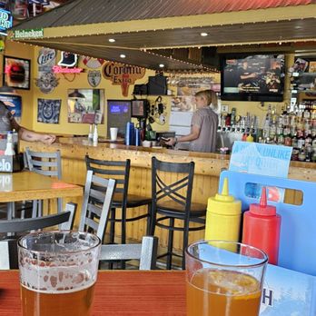 3rd Base Bar Grill - Cold beer with a lot is selections. - The image shows two glasses of beer on a wooden table inside a bar. The background features a bar counter with bottles, a bartender, and various colorful signs and decorations, including a neon sign that reads 'COLD BEER WINE'. The ceiling has wooden beams, and there are chairs and tables arranged for patrons. The setting appears lively and casual, with a variety of signage and decor typical of a bar environment. - Burgers Near Me - La Porte, Indiana