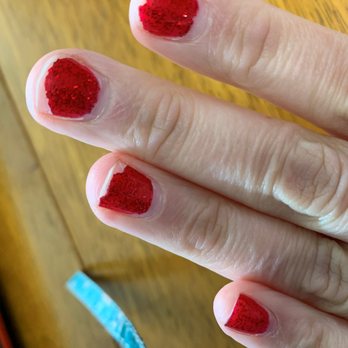 Serenity Nail Spa - 10 Days after no-chip manicure - A close-up image shows a hand with four fingers, each nail painted with red glitter nail polish. The nails are short and well-manicured, with the polish evenly applied. The background features a wooden surface, and part of a blue object is visible in the lower left corner. The lighting highlights the sparkle of the glitter on the nails. - Nail Salons Near Me - Franklin, Wisconsin