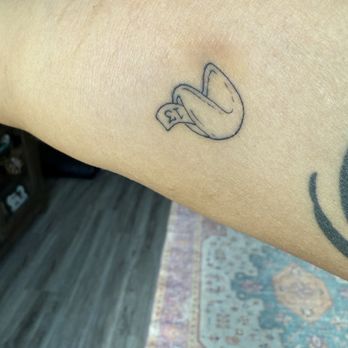 Ink Couture - A tattoo depicting a hand holding a dollar bill with the number 13 is on a person's forearm. The tattoo is outlined in black ink and appears to be a simple, cartoon-style design. The person's skin is visible, and part of another tattoo can be seen on the upper arm. The background shows a room with a wooden floor and a patterned rug. - Tattoo Near Me - Austin, Texas