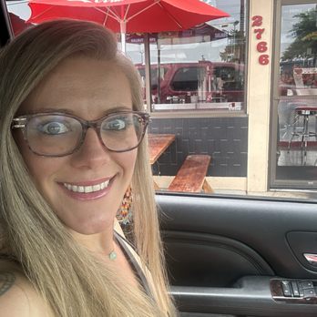 Toxic Burger - Waiting for the grub. - A woman with long blonde hair and glasses is smiling in a selfie taken inside a vehicle. She is wearing earrings and a necklace. Through the car window, a restaurant with a red Coca-Cola umbrella, outdoor wooden tables, and benches is visible. The restaurant's exterior features a sign with the address 2766 and large windows showing parked cars and a street scene. The image captures a casual moment with the woman enjoying her time near the restaurant's outdoor seating area. - Chicken Wings Near Me - Eugene, Oregon