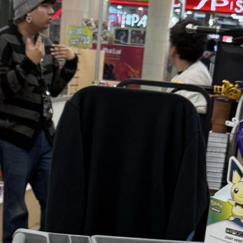 Entertain Mart - These two stood and gossiped for a good 15-20 minutes. Ignoring customers. Yes I timed it while being ignored. - The image shows two people in a retail store, possibly a gaming or electronics shop, engaged in conversation. One person, wearing a gray beanie and earrings, stands with hands near their chest, while the other, with dark hair, is seated and facing them. The background includes store signage, shelves with products, and a display case with items and prices. The setting features bright lighting and colorful interior decor. - Video Game Stores Near Me - Aurora, Colorado