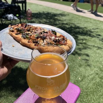 Frontyard Brewing - Belgium Golden Ale and Prosciutto pizza - The image depicts a person holding a plate with a slice of pizza topped with various ingredients and a glass of beer on a pink table. The background shows an outdoor area with grass, a paved walkway, and several people standing and walking, suggesting a social gathering at a brewery or outdoor event. - Breweries Near Me - Spicewood, Texas