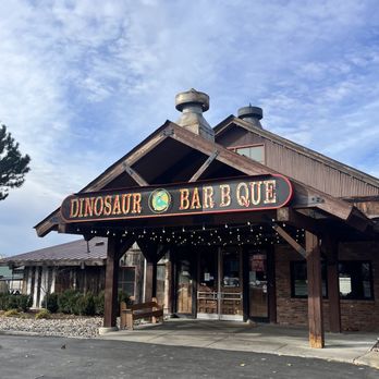 Dinosaur Bar-B-Que - The photo depicts the exterior of Dinosaur Bar-B-Que, a restaurant with a rustic wooden facade and a prominent sign displaying its name. The sign reads 'DINOSAUR BAR-B-QUE' with a dinosaur logo in the center. String lights are hung beneath the sign, and there is a bench near the entrance. The building has a sloped roof with visible chimneys, and the surrounding area includes a paved parking lot and some landscaping with bushes. The sky above is partly cloudy. - Barbeque Near Me - Troy, New York