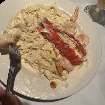 Casa Capitano - Seafood Alfredo - A white plate holds seafood Alfredo with wide pasta noodles coated in a creamy white sauce. The dish includes lobster, shrimp, and possibly other seafood, garnished with small green herbs. The plate is on a white tablecloth, with a fork held by a person in the foreground and a hand visible at the top. A glass of beverage is partially visible in the background. - Italian Near Me - Rouses Point, New York