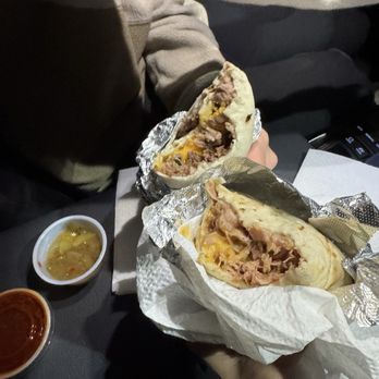 Perico's Albuquerque - A person is holding two beef and cheese burritos wrapped in foil and paper, with one burrito partially unwrapped to show beef, cheese, and other fillings. The burritos are placed on a dark surface with side sauces and a small bowl of soup nearby. The setting appears to be inside a vehicle, with a seat and part of a dashboard visible. - Mexican Near Me - Albuquerque, New Mexico