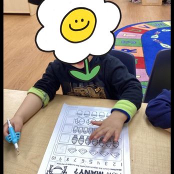 Lightbridge Academy - A child is seated at a table, working on a worksheet with a pen, in a classroom setting. The child's face is covered with a smiley flower sticker. - Child Care & Day Care Near Me - Millburn, New Jersey