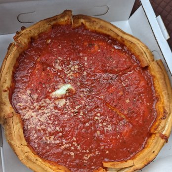 Cipriani's Pizza - A deep dish pizza with a thick, golden-brown crust is shown inside a cardboard pizza box. The pizza is topped with a layer of tomato sauce and melted cheese, with some cheese sprinkled on top. The pizza appears to be freshly baked, with a slightly uneven crust and a rich red sauce. The box is partially open, revealing the pizza inside, and the background includes a textured surface. - Pizza Near Me - Chicago, Illinois