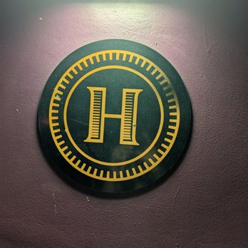Highball - A round sign featuring a yellow 'H' in the center, mounted on a purple wall, with a light source above illuminating the area. - Cocktail Bars Near Me - Phoenix, Arizona