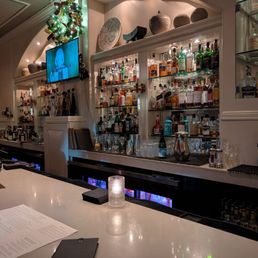 A bar with liquor bottles, glassware, and decorative pottery on shelves, illuminated by spotlights, with a white countertop and a small candle.