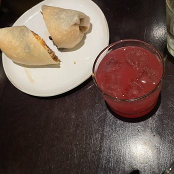 The Roost - Two spring rolls are placed on a white oval plate, with one cut open to reveal the filling. Next to the plate, there is a short glass filled with a red, ice-filled drink, and a clear glass of water. The setting is on a dark wooden table, likely in a restaurant or dining environment. - Coffee & Tea Near Me - Washington, DC, District of Columbia