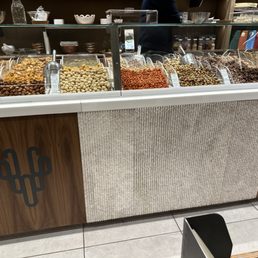 A nice display of fruits and nuts - A shop counter features a glass display case containing an assortment of nuts and dried fruits, each in separate containers with scoops. The counter has a wooden panel with a cactus logo and a textured beige panel below. The background shows shelves with bowls, jars, and books, indicating a retail environment for Middle Eastern or gourmet foods.