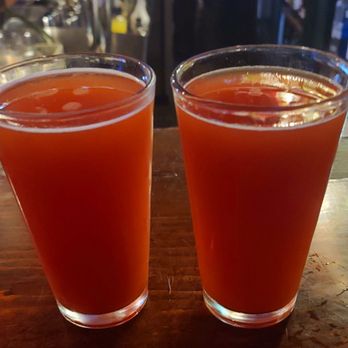 Murphys Historic Hotel - Hair of the Dog - Two glasses filled with a reddish-orange beverage are placed on a wooden bar counter, with a blurred bar background. - Hotels Near Me - Murphys, California