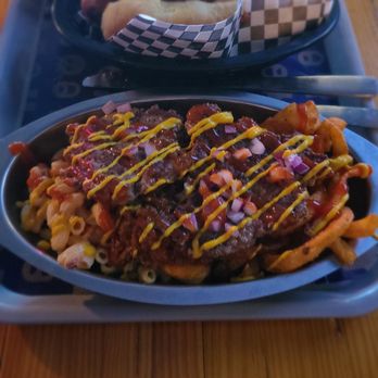 The Beer Keep - Close up of their plate - The image shows two food dishes on a tray. The front dish contains loaded fries topped with meat, cheese, diced onions, and mustard, with additional sauces. The background dish features a hot dog in a bun, topped with mustard and other condiments. Both dishes are served on black plastic trays, placed on a wooden table, with utensils nearby. - Beer Bar Near Me - Buffalo, New York