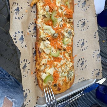 George's Local - Buffalo Chicken Flat Bread - A rectangular flatbread pizza topped with melted cheese, shredded carrots, sliced celery, and chunks of chicken. The pizza is served on branded paper from George's Local, with a side of fried item at the top left corner. The tray is placed on a metal outdoor table, with a fork resting on the tray and a person's ripped jeans visible in the bottom left corner. - New American Near Me - Cedar Falls, Iowa