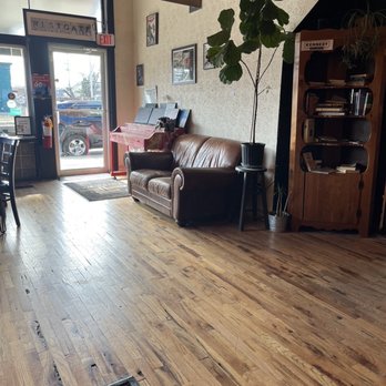 Third Way Cafe - Inside - The image shows the interior of a coffee shop with wooden flooring and a leather sofa positioned against a wall. A large potted plant is placed beside the sofa on a small table. The wall behind the sofa is decorated with framed artwork. Near the entrance door, there is a red piano and a music stand. The door has a window, and outside, a parked car is visible. The space appears cozy with natural light coming through the window. - Coffee & Tea Near Me - Columbus, Ohio