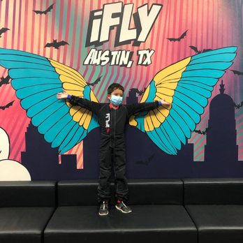 iFLY - A child wearing a face mask and a black jumpsuit stands with arms extended, mimicking butterfly wings painted on a colorful mural at iFLY indoor skydiving facility in Austin, TX. The mural features large blue and yellow butterfly wings, with a background of pink and purple stripes, silhouettes of bats, and cityscape elements. The child is positioned on a black bench in front of the mural, creating the illusion of having butterfly wings. - Skydiving Near Me - Austin, Texas