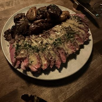 Pretty Penny - Steak and fingerling potatoes - A plate features sliced steak topped with a creamy herb sauce, accompanied by roasted fingerling potatoes and a portion of roasted mushrooms, set on a wooden table. - Cocktail Bars Near Me - Phoenix, Arizona