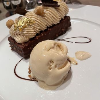 Daphne - A dessert plate displays a rectangular chocolate cake with a glossy finish, topped with piped cream, edible gold leaf, and chocolate garnishes. A scoop of vanilla ice cream is placed beside the cake, with a drizzle of chocolate sauce on the plate. The background includes wine glasses and a blurred dining setting. - Seafood Near Me - Toronto, Ontario