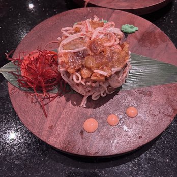 Mamasushi - Kani Salad - A sushi dish featuring a mound of noodles topped with a mixture of cooked ingredients, garnished with a green wasabi dollop and red shredded garnish, served on a round wooden plate with a leaf decoration. The plate is placed on a dark, speckled surface. - Sushi Bars Near Me - Brooklyn, New York