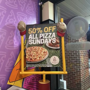Mazala Pizza - Sundays - A freestanding signboard outside Mazala Pizza displays a promotion for 50% off all pizza on Sundays. The sign features a large image of a pizza and a smaller image of a plate of chicken wings. Two beige and red caps hang on the top corners of the sign. The sign is positioned near a glass window and brick wall, with part of the interior and exterior environment visible. The sign includes terms about the offer being valid only on Sundays until the completion of football playoff games. - Chicken Wings Near Me - Naperville, Illinois