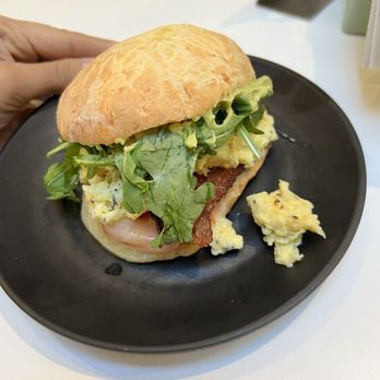 Bōm Dough - good egg - A breakfast sandwich featuring scrambled eggs, leafy greens, bacon, and a bread roll, served on a black plate at a cafe. - Brazilian Near Me - Cambridge, Massachusetts