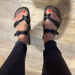 Pedicure - A photograph taken from above shows a person wearing black sandals with adjustable straps, standing on a wooden floor with a textured surface. The person's toenails are painted red, and they are wearing black pants that reach just above the ankles. The image captures the feet, sandals, and part of the legs, with the background consisting of a wood-patterned floor.