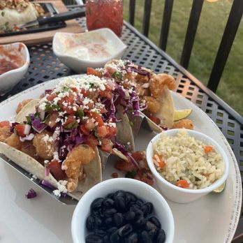 Sol Agave - Bang bang shrimp tacos - A plate of two fish tacos with shredded cheese, vegetables, and toppings, accompanied by rice and black beans, on an outdoor dining table. - Mexican Near Me - Grimes, Iowa