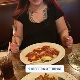 Cheese ravioli and bolognese ! - A woman with long brown hair, tattoos, and a black shirt is seated at a restaurant table, smiling with a fork and knife, with two plates of pasta and a drink.