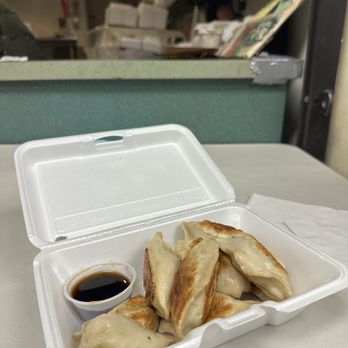 Dumpling King - Pan Fried Pork and Leek Dumpling - A white foam takeout container is open, revealing several fried dumplings with a golden-brown exterior and a small cup of soy sauce. The container is placed on a table in a casual environment, with a background showing a cluttered counter and shelves with various items. - Dim Sum Near Me - Boston, Massachusetts