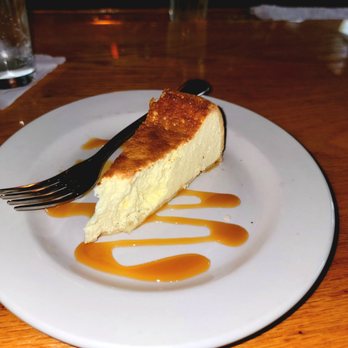 Pasquale Rigoletto Restaurant - Just a taste of the ricotta cheesecake - thank you! - A slice of ricotta cheesecake with a slightly browned top is placed on a white round plate. The cheesecake has a creamy texture and is garnished with a drizzle of caramel sauce underneath. A black fork rests on the plate, and the background shows a wooden table with some glasses and napkins, indicating a dining setting at Pasquale Rigoletto Restaurant. - Italian Near Me - Bronx, New York