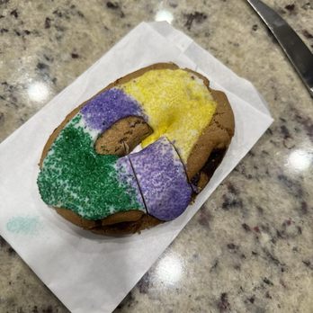 BATTER - King cake cookie - A cookie decorated with yellow, purple, and green icing and sprinkles, resting on a piece of paper on a granite countertop. - Desserts Near Me - Hammond, Louisiana