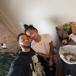 A man and woman pose for a selfie at a table in an indoor setting, with a white wall and some plants in the background.