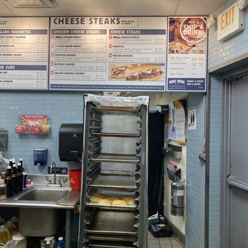 Jersey Mike's Subs - Menu - A metal bakery rack with several trays of bread is positioned in a restaurant kitchen, with a menu board above. - Sandwiches Near Me - Warwick, Rhode Island