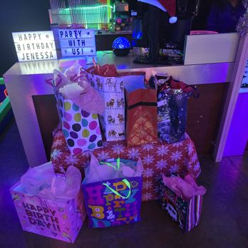 Lunar Golf - Gift bags with tissue paper are placed on a table covered with a red snowflake-patterned cloth at Lunar Golf in Albuquerque, NM. The scene includes birthday signs, one reading 'HAPPY BIRTHDAY JENESSA' and another 'PARTY WITH US!', illuminated by colorful, neon-like lighting. The setting appears to be a birthday celebration within the indoor mini-golf venue, with festive decorations and a sign about Lunar Golf's promotional offer. - Mini Golf Near Me - Albuquerque, New Mexico