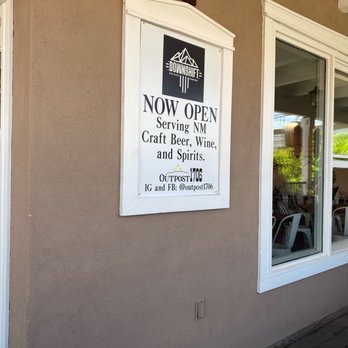 Downshift Brewing: Outpost 1706 - Cute indoor outdoor bar off the Square - A vertical signboard mounted on a beige exterior wall of a building features the logo and name of Downshift Brewing. The sign indicates that the brewery is now open and serves craft beer, wine, and spirits. The sign also includes social media handles and contact information. A window with a white frame is visible nearby, showing part of the interior with chairs and tables. The setting appears to be in a commercial or retail area. - Beer, Wine & Spirits Near Me - Albuquerque, New Mexico