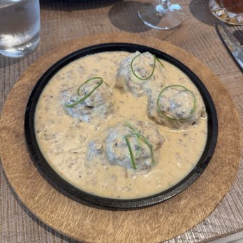 Rumis Kitchen - Za'atar Biscuits & Gravy - Four meatballs in a creamy, herb-infused sauce are presented on a black skillet, garnished with thin green onion slices, on a wooden serving board. - Persian/Iranian Near Me - Houston, Texas